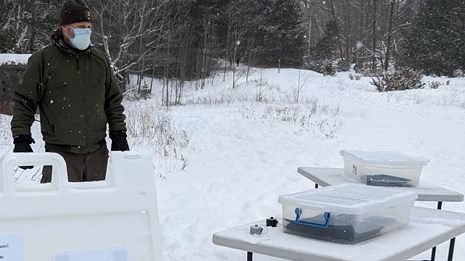 A ranger stands in the snow in front of a table and information board.