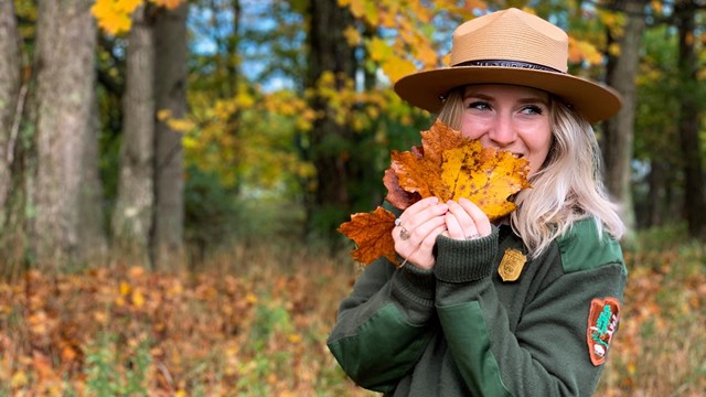 A female park ranger in uniforms holds maple leaves in front of her face.