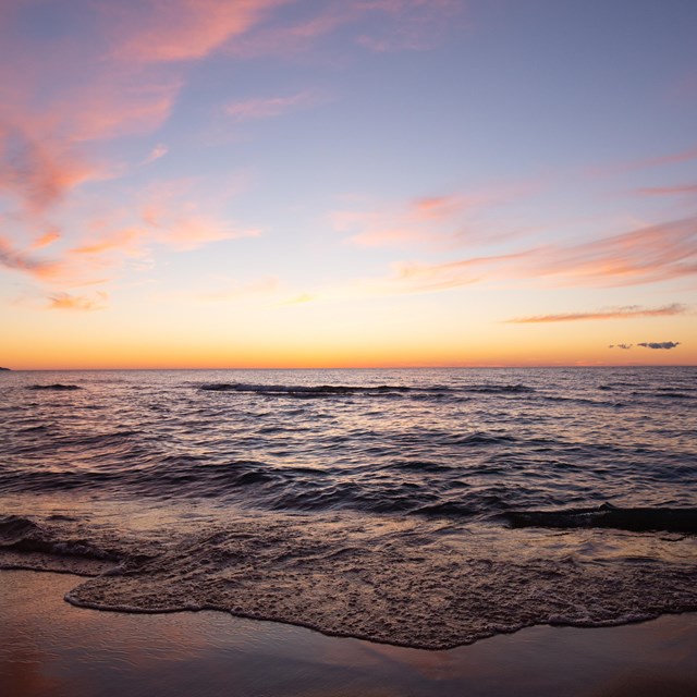 A sun sets along the shores of a large body of water and sandy beach landscape.