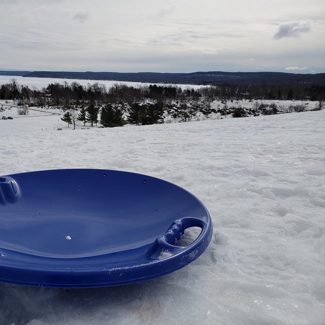 A blue saucer sled sits atop a snowy hill with bare trees in the background.