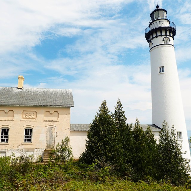 Looking up from the ground, a faded yellow building is dwarfed by the towering white lighthouse. 