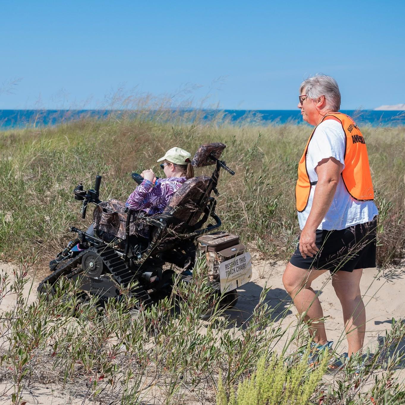 A person in a track wheelchair on a sandy beach with another person walking behind