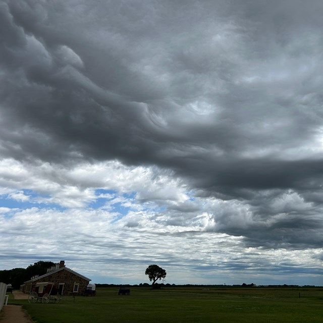 Dark storm clouds over a grasslands landscape with a small building in the lower left.