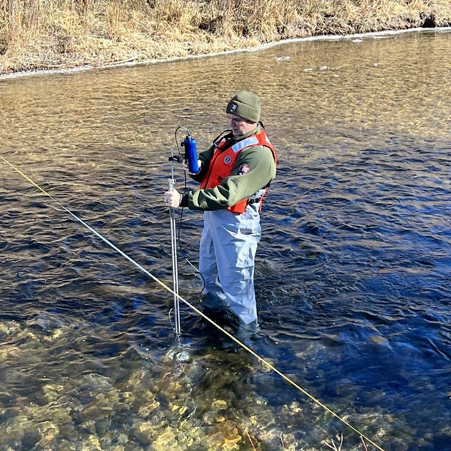 A scientist standing in a stream holding equipment that measures water flow.