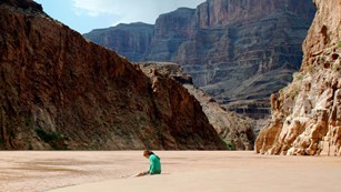 A woman sits on the sandy shore of a riverbank in a wilderness mountain canyon.