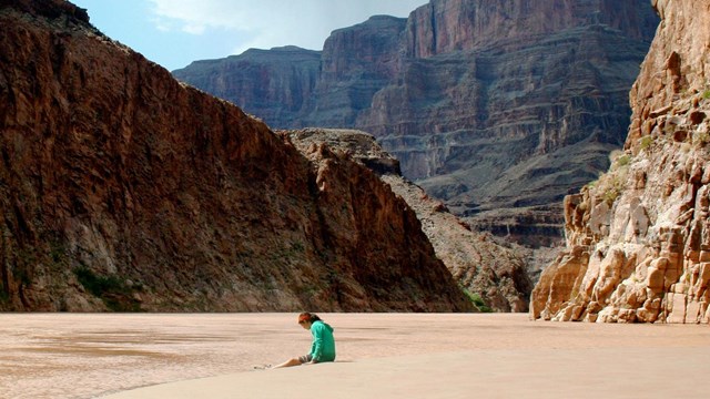 A woman sits on the sandy shore of a riverbank in a wilderness mountain canyon.