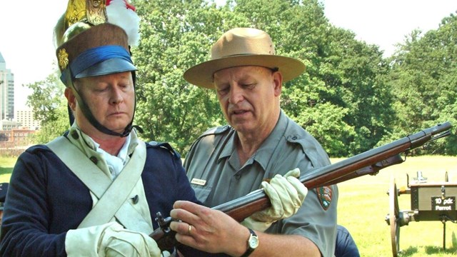 A park ranger and a man in an 1800s military uniform hold a rifle.