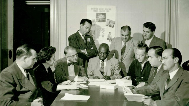 Nine men and one woman sitting and standing around a table looking at documents.