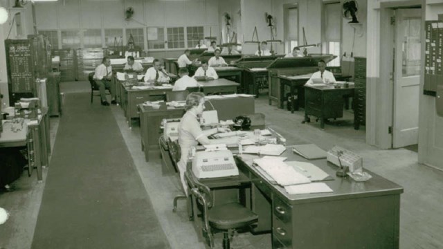 A woman smiles at the camera while sitting at a drafting table. 