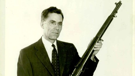 A black and white photo of a man in a suit holding the M1 Rifle. 