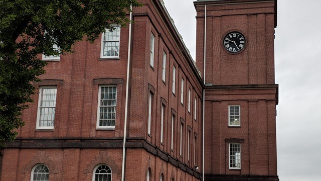 Three story brick building with six story clock tower in front.  American flag waving from tower.