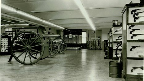 A black and white photo of a museum floor with display cased with firearms and cannons. 