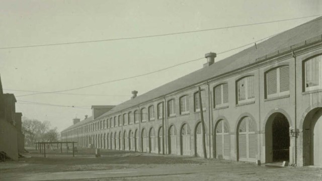 A black and white photo of a long building with multiple windows and a small house to the left. 
