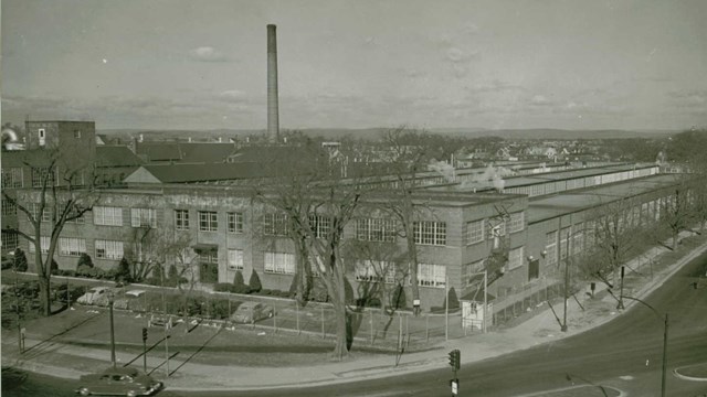 A black and white photo of a multistory building with a smoke stack in the background. 
