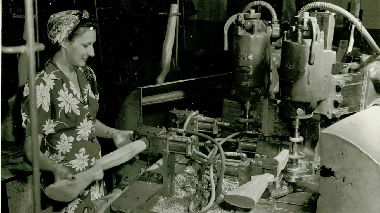 A black and white photo of a women in an dress and apron working on a gun stock. 