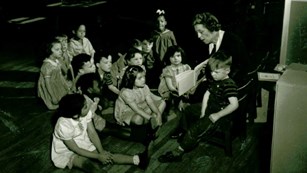 A black and white photo of a teacher sitting with young children reading them a book. 