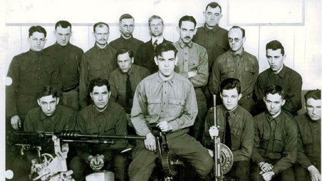 Men look at the camera.  Machine guns are laid out in front of them.