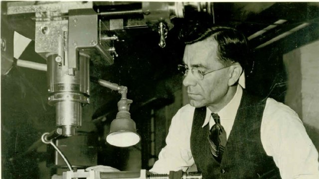 A black and white photo of a man with glasses in a suit, standing at a machine making firearm parts.