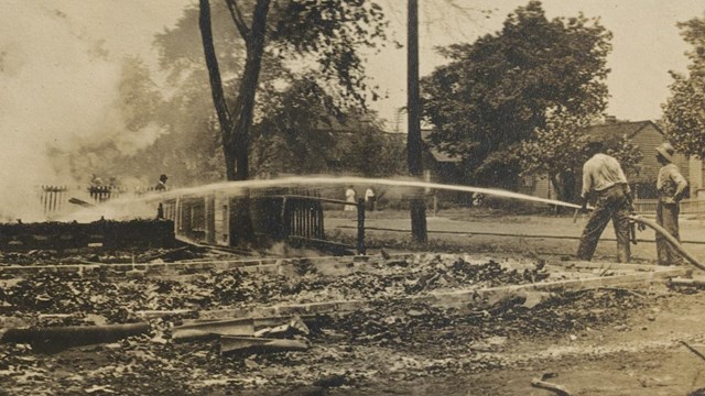 Historic photo of two African American firefighters using a hose to extinguish a burning house.