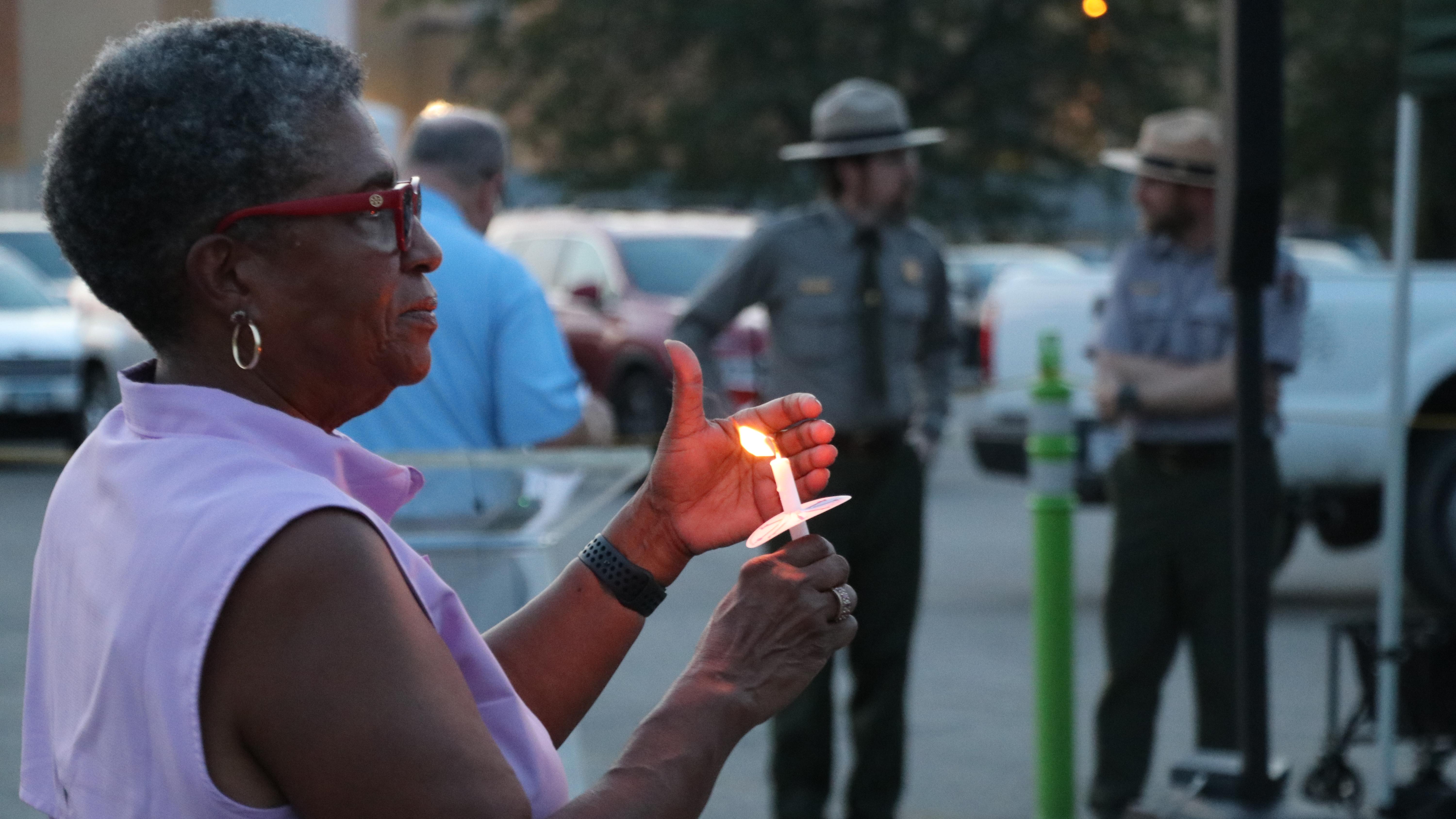 A woman holding and shielding the flame on a burning candle, with a crowd in the distance.