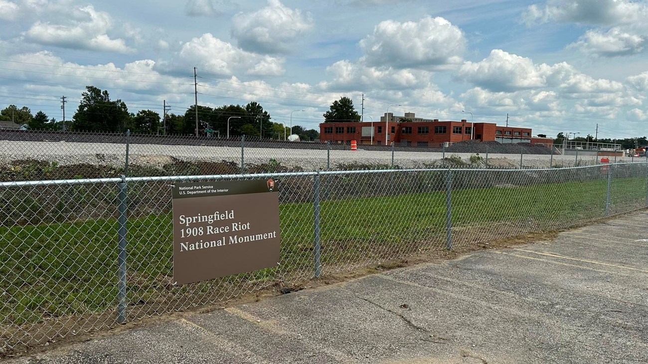 A national park entrance sign hangs on a chain link fence at the edge of a paved parking lot.