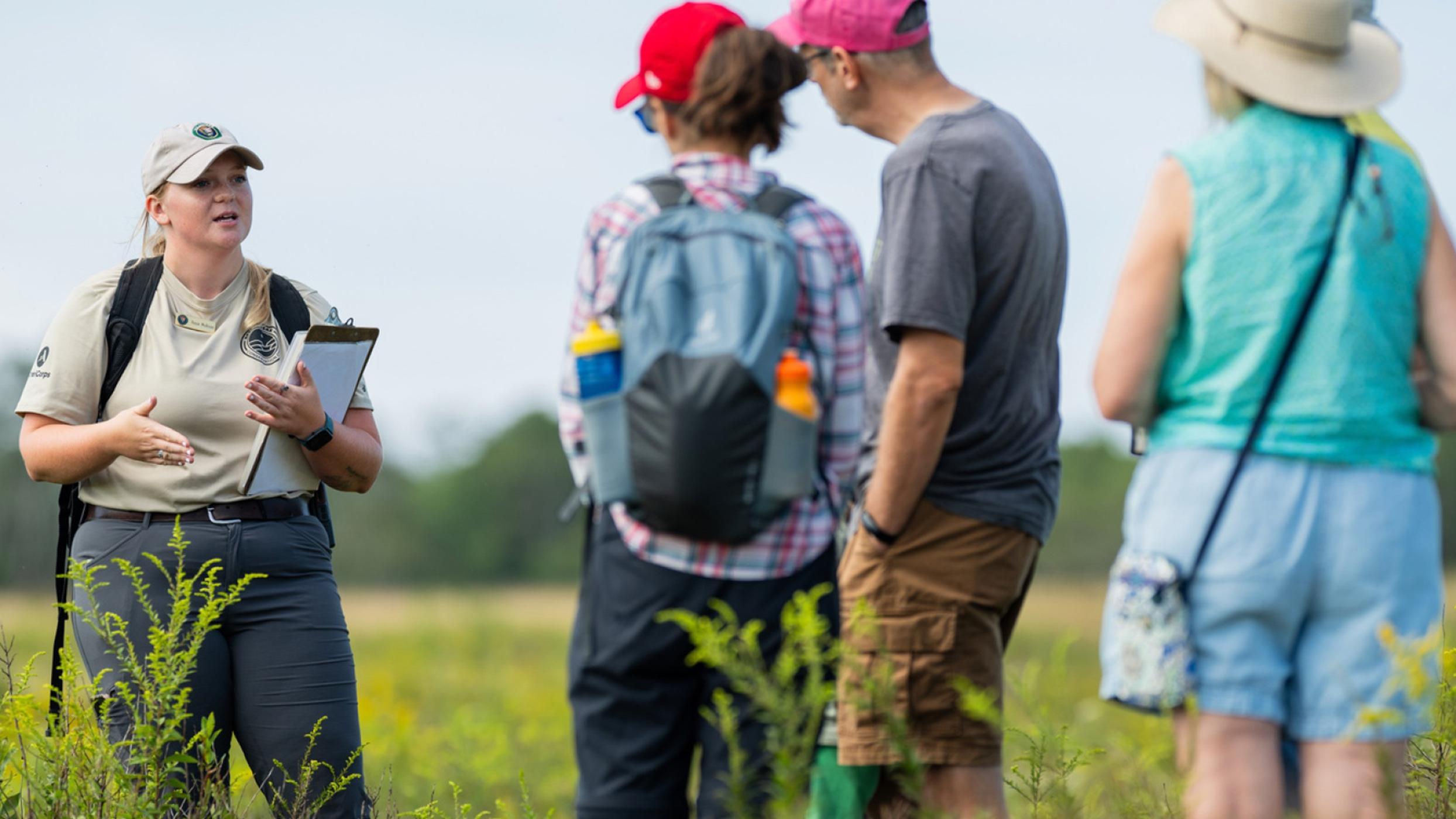 A woman in tan uniform holding a clipbord talkng to two visitors in the middle of a grassy field.
