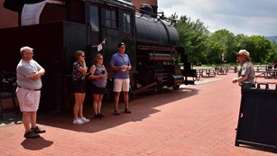 A socially distanced crowd stands outside in front of a black train as a ranger speaks with them