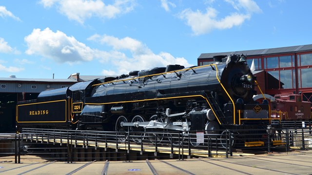 Large, black, steam engine outside on turntable. Blue skies with sporadic clouds