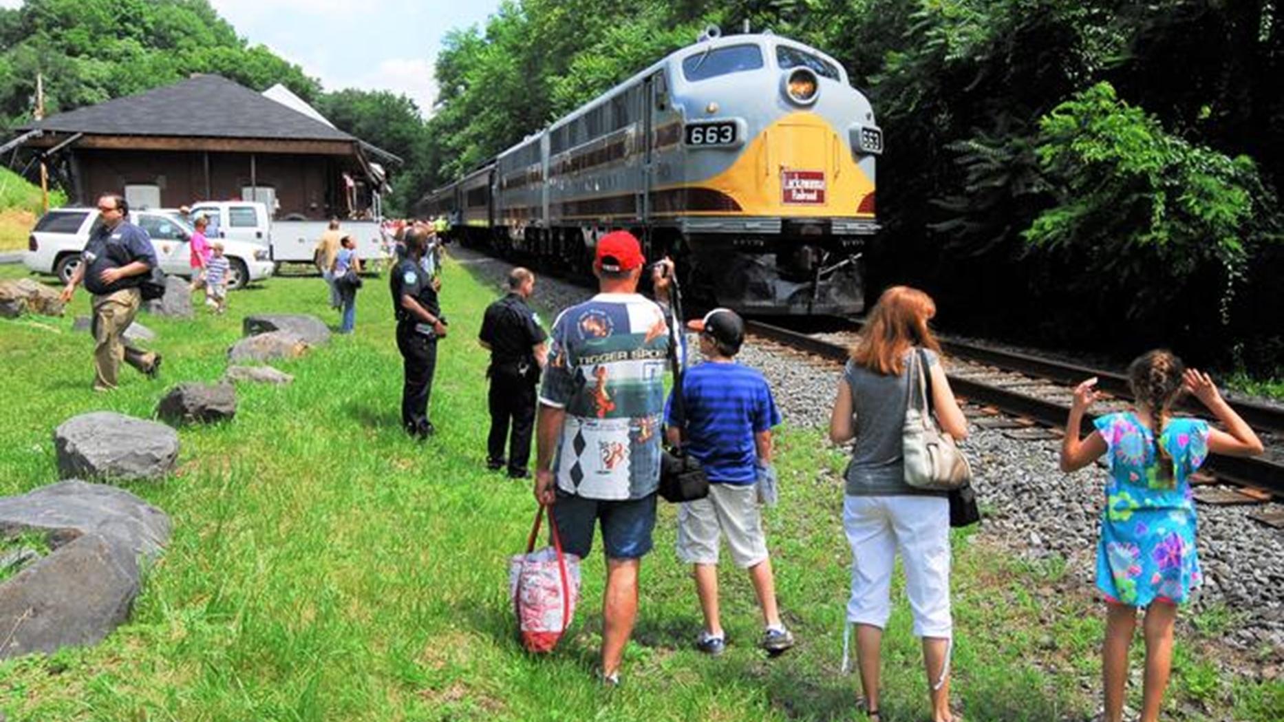 Diesel Locomotive in gray, gold, and red paint on a track next to a depot building and a small crowd