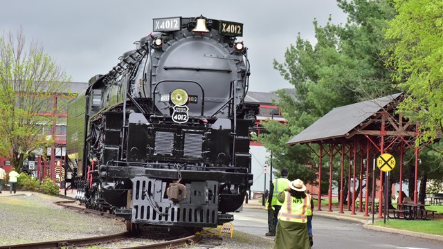 Large black train stationed on a curved track with a park ranger in the foreground