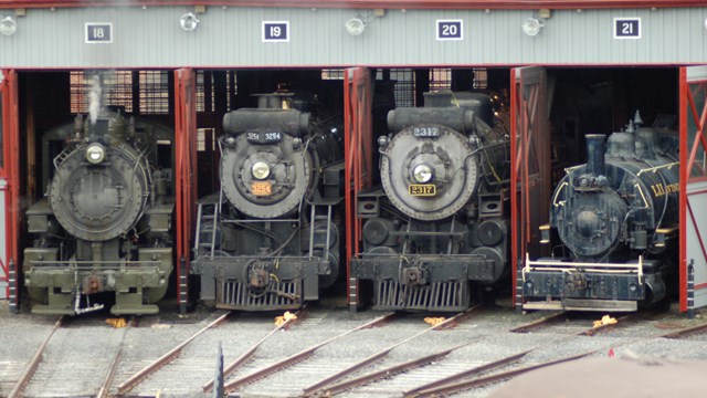 Four steam locomotives sitting in the open doors of a round house. 