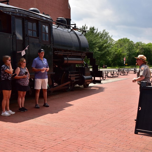 A socially distanced crowd stands outside in front of a black train as a ranger speaks with them