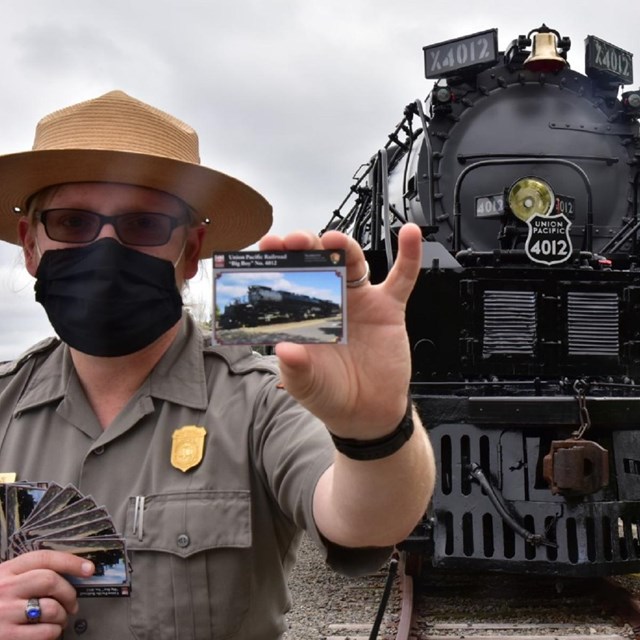 Park ranger wearing a mask and holding a trading card in front of Big Boy train