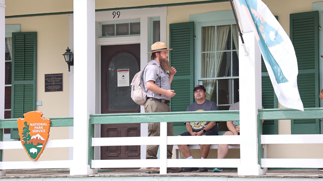 Park Ranger stands on the porch of the Jean Baptiste Valle house talking to a group of visitors.
