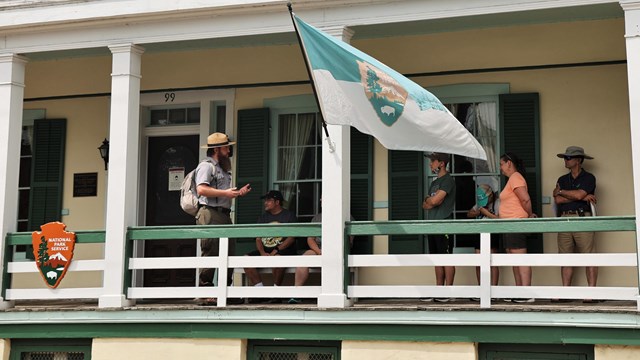 Park ranger talking to a group of people on a covered porch or a cream colored house.