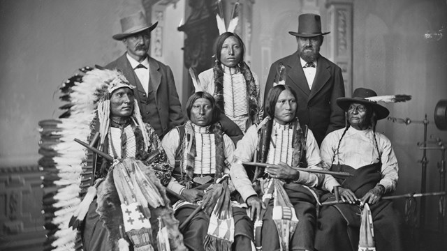 Portrait of five members of the Arapaho Nation in traditional clothing and two men in suits.