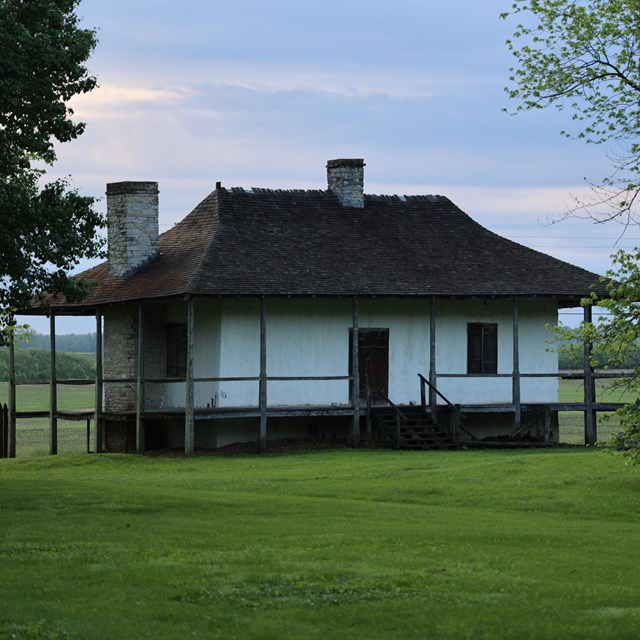 Sun setting on white home with brown accents, sitting in a grassy field with trees in front of it