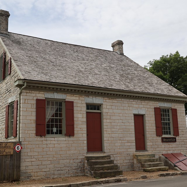 Light tan stone building with red doors and shutters and a brown roof.