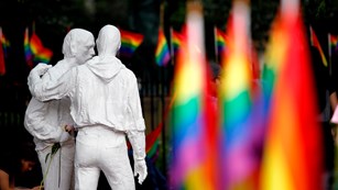 Life sized statues of two men standing inside Christopher Park, surrounded by pride (rainbow) flags.