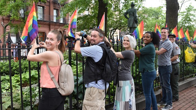 Five visitors and one park ranger standing next to an iron fence as they post rainbow flags.