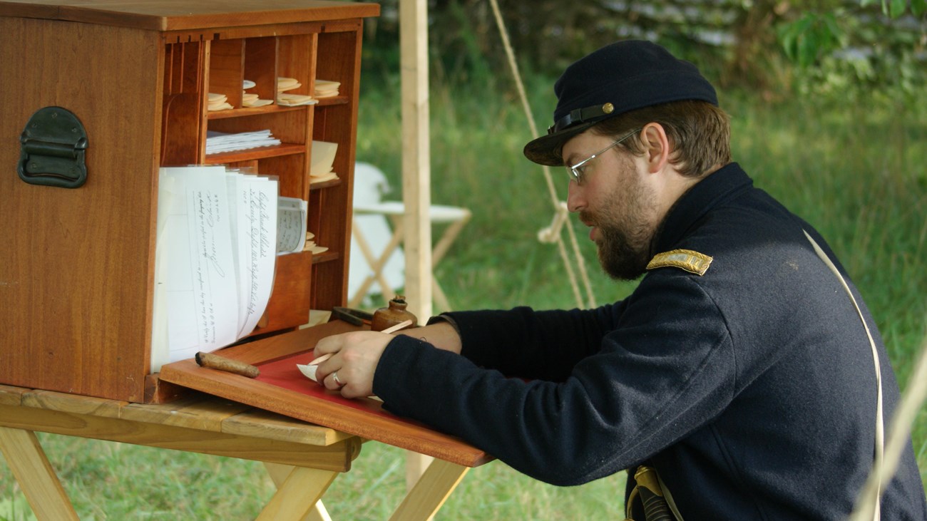 A living historian dressed as a Union officer writes on paper at a field desk.