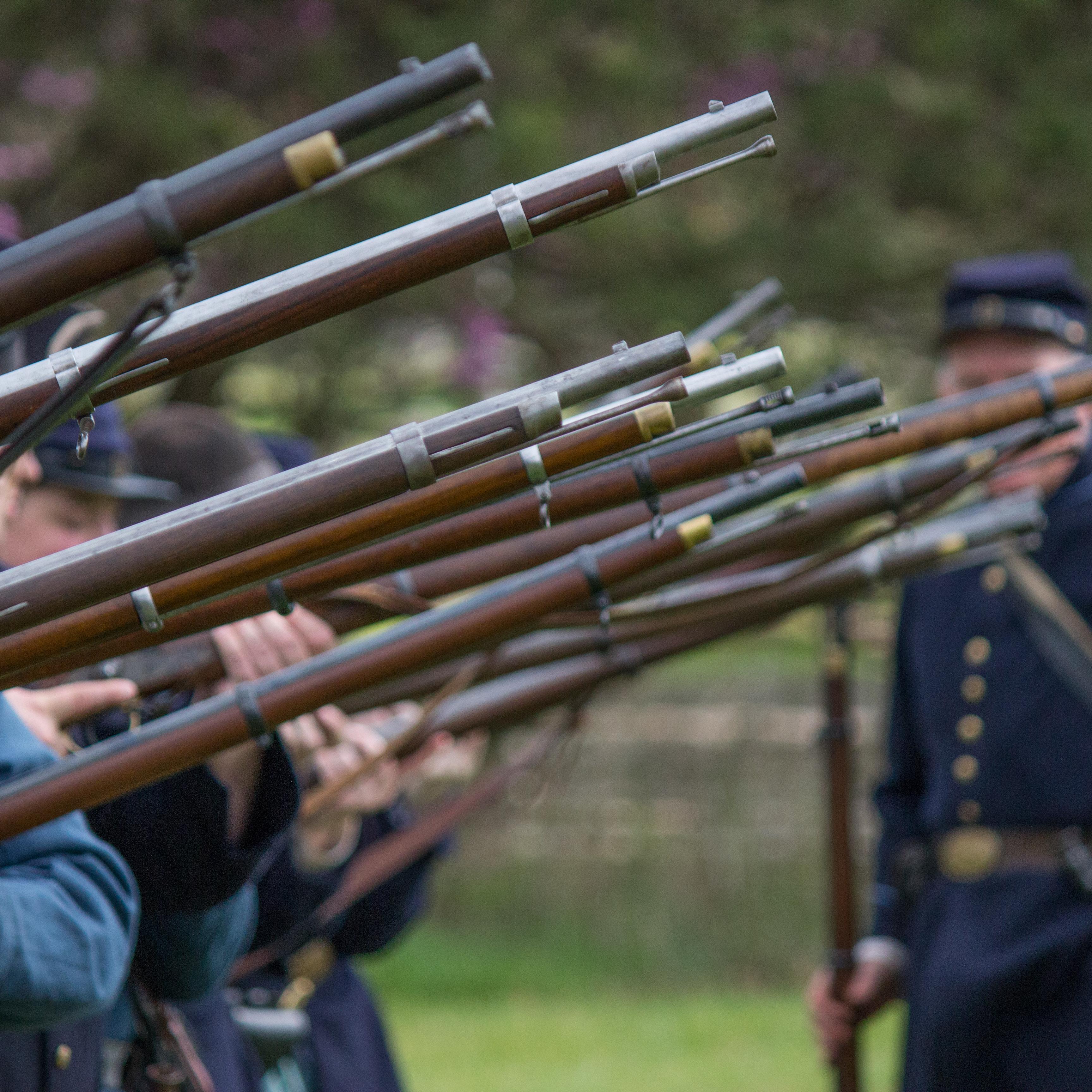 A row of soldiers hold muskets in the air with a Union soldier looking on in the background.