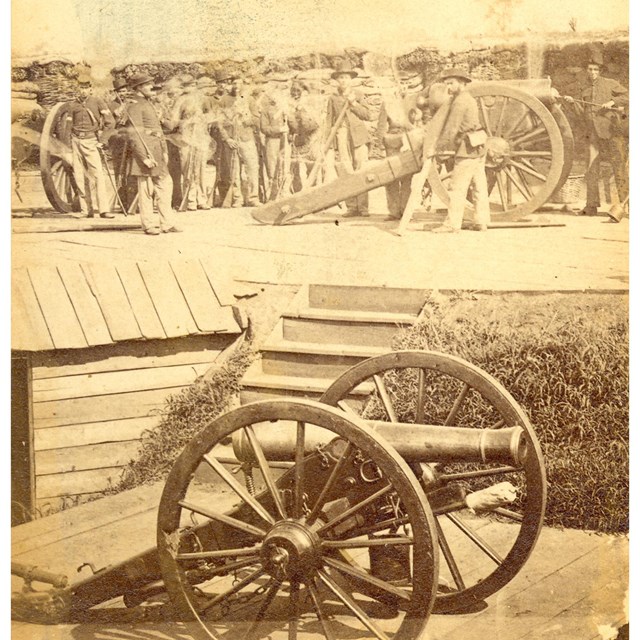 A black and white photo of a cannon on a wooden platform, with soldiers and another cannon on a plat