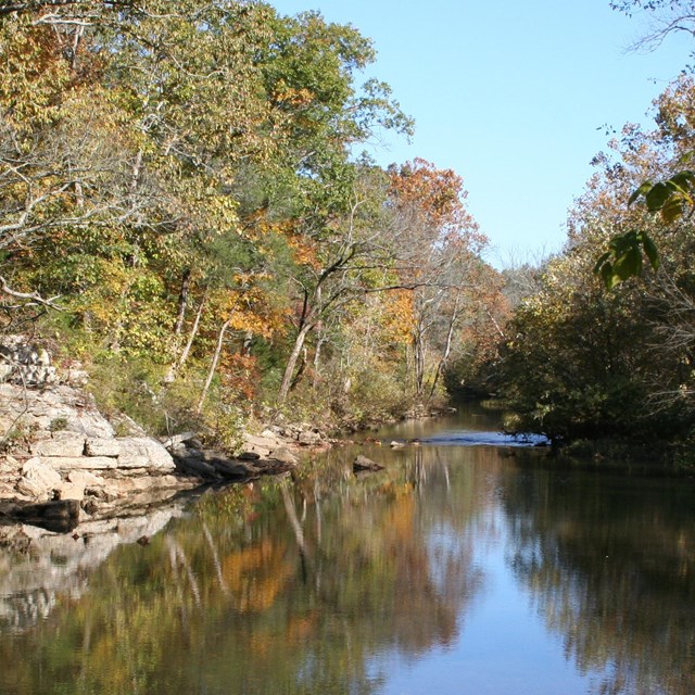 A still river with rocks along the bank and trees covering both banks. Text of the reverse side is s