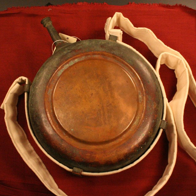 A copper-colored Civil War canteen and white cloth sling on a red felt background
