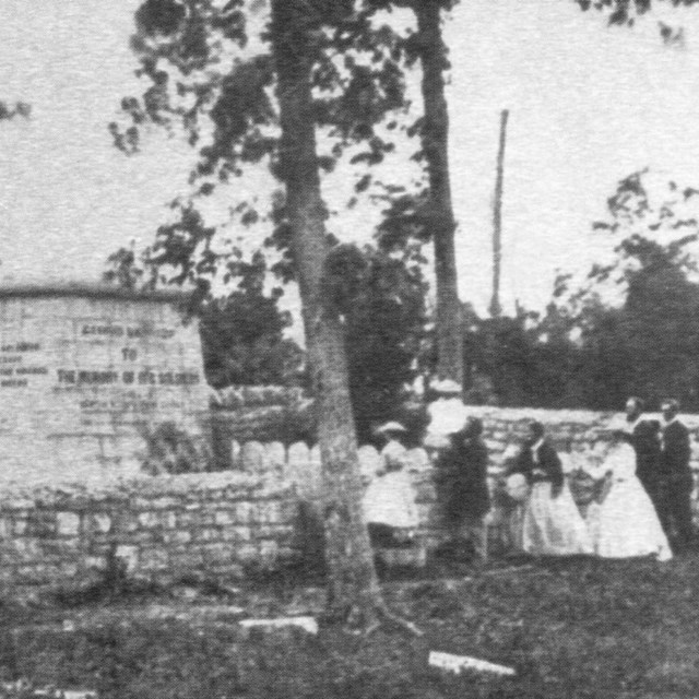 A black and white photo shows a cubed stone structure with a stone wall around it.