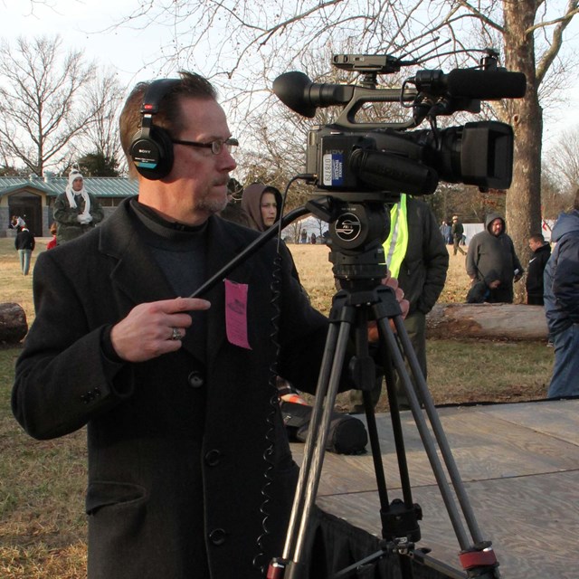 A man in a black coat looks through a video camera.