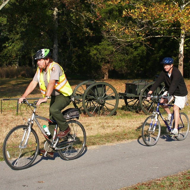 A park ranger wearing a bike helmet rides a bike in front of a bicyclist in shorts.