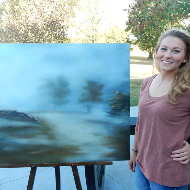 A young woman stands next to her painting of the battlefield.