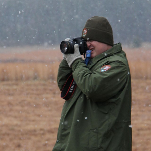 A man in a green jacket and knit cap takes a photograph in a field.